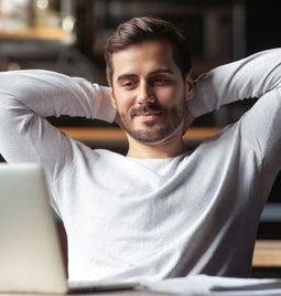 Man with arms behind his head relaxing as he looks at a laptop