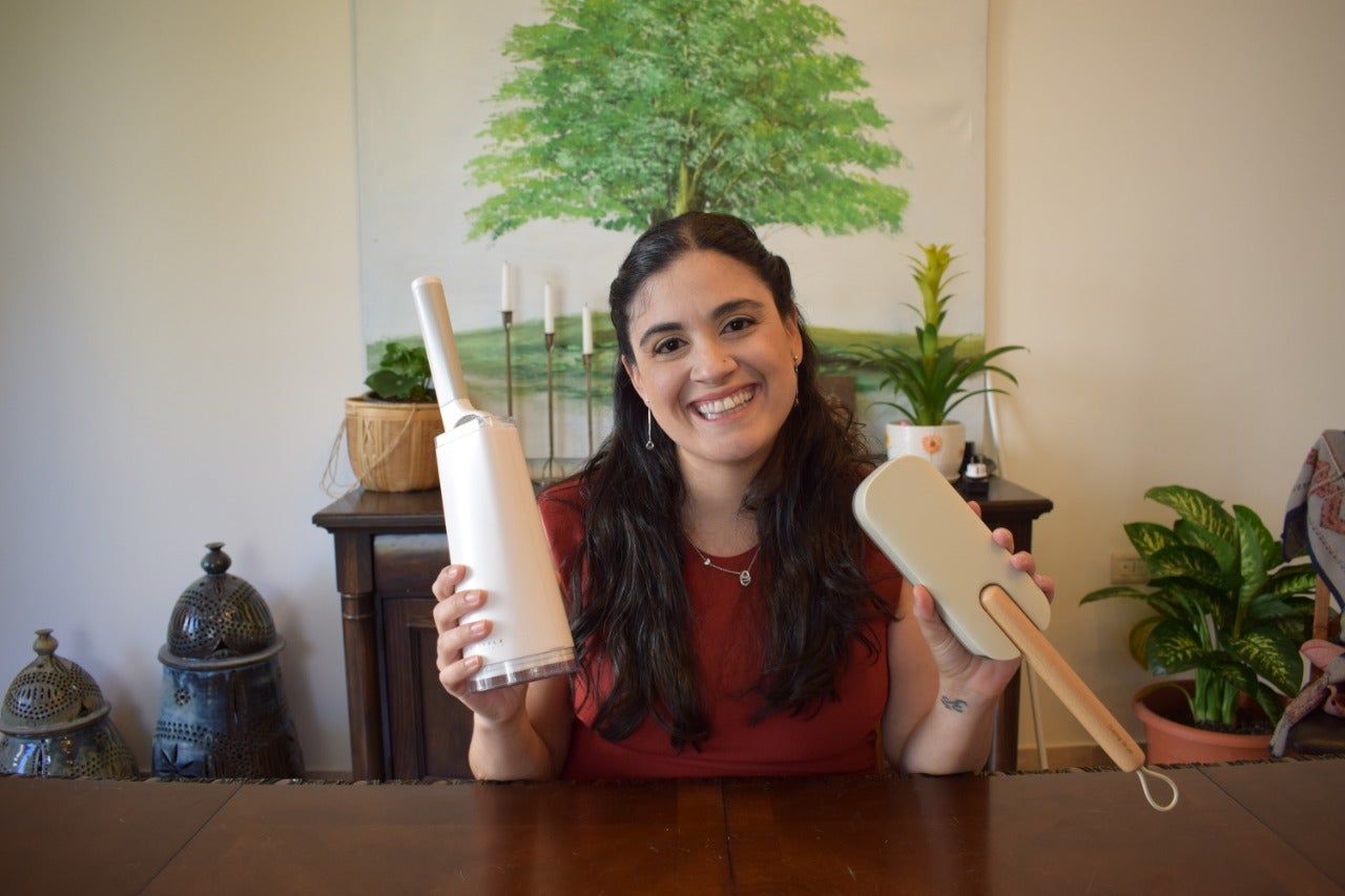 headshot (1) waist high headshot of woman holding two lint brushes smiling in front of wood table with tree painting behind