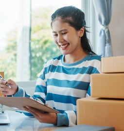 Woman stands behind a pile of package boxes smiling at her clipboard