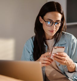 woman sits with an open laptop looking at her phone in her hand