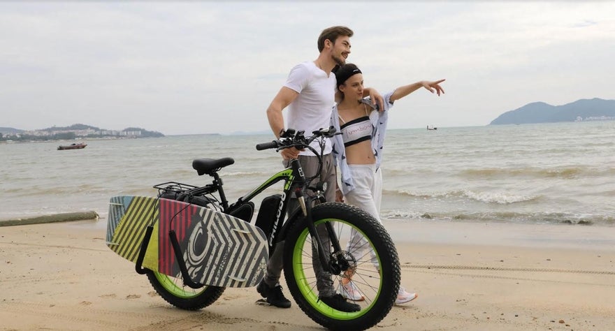 Ovo Bikes Promo 2 A man and woman walking along the beach. The man has his arm around the woman and is steering his bike with the other. The woman is leaning into him and pointing into the distance.