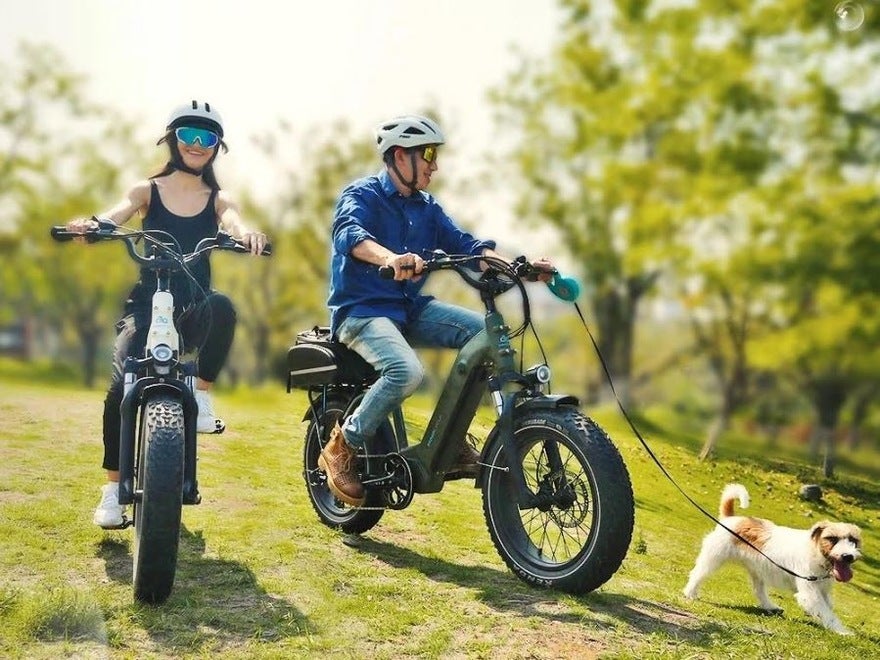 Ovo Bikes Promo A man and a woman on electric bikes, riding across a field with trees in the background. The man is holding a dog on a lead.