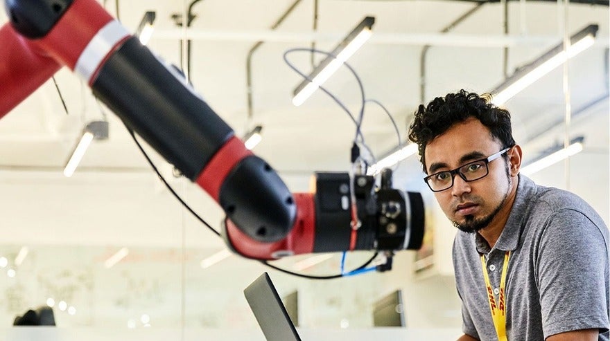 A man with glasses attentively observes a red and black robotic arm in a lab setting, with a laptop in front of him and lighting fixtures overhead.