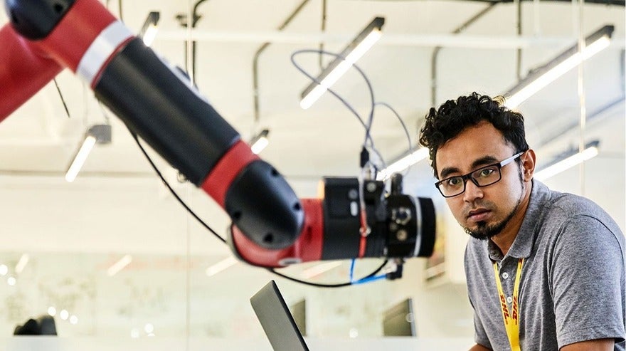 DHL A man with glasses attentively observes a red and black robotic arm in a lab setting, with a laptop in front of him and lighting fixtures overhead.