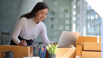 Female Entrepreneur looks at her laptop while making notes surrounded by order boxes