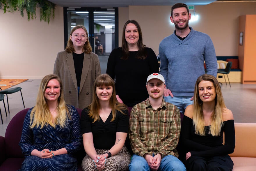 Website Builder Expert Team Photo Seven members of the WBE team smile at the camera in an office