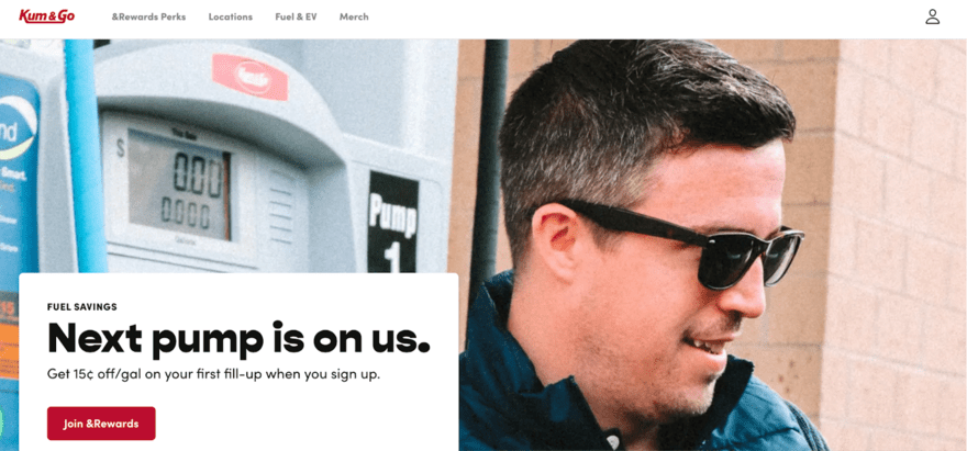 A man in sunglasses smiles at a fuel station next to a pump displaying zero cost, under a promotional sign offering fuel savings for new members.