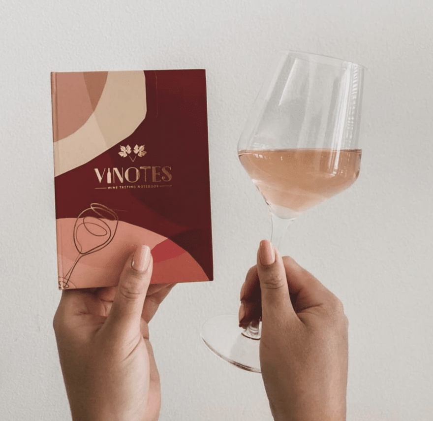 Screen Shot 2023-02-15 at 10.27.51 AM (1) (1) Hands holding a glass of rose wine in one hand and a notebook in the other